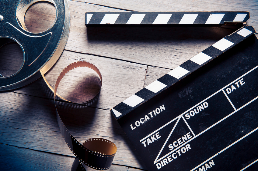 A clapperboard and film reel are positioned on a wooden table. The film reel is a light metal and is partially unspooled. Next to it is a black-and-white film slate with the clapper opened. There is nothing written on the clapperboard.