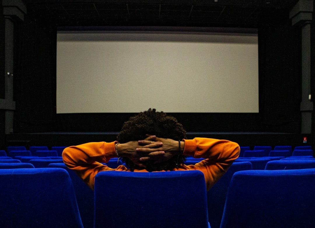 Person seated in a cinema, facing a blank screen with hands behind their head